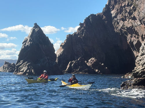 Two people kayaking on blue water in front of large dark rocky cliffs under a blue sky with white clouds. Ingonish Island.