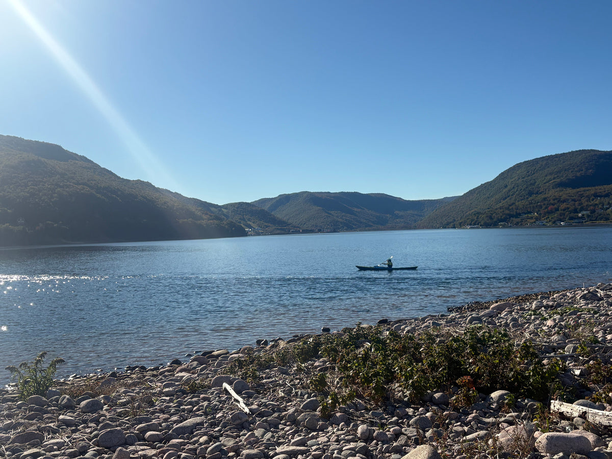 A person kayaking in a harbour framed by rocky shores in the foreground and green hills under a clear blue sky.